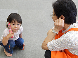写真：おもちゃの携帯電話を耳にあてる女児を見守る、オレンジ色のエプロンの女性