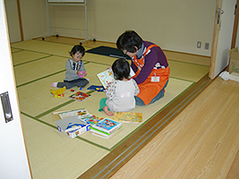 畳の部屋で、おもちゃで遊ぶ女児と、別の子に絵本を見せるエプロンの女性