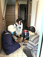 写真：玄関で座って靴に手を当てる男児と、かがんで見守る3人の大人