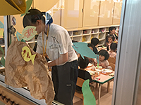 写真：窓のそばで作業する生徒と、その後ろで食事する園児たち